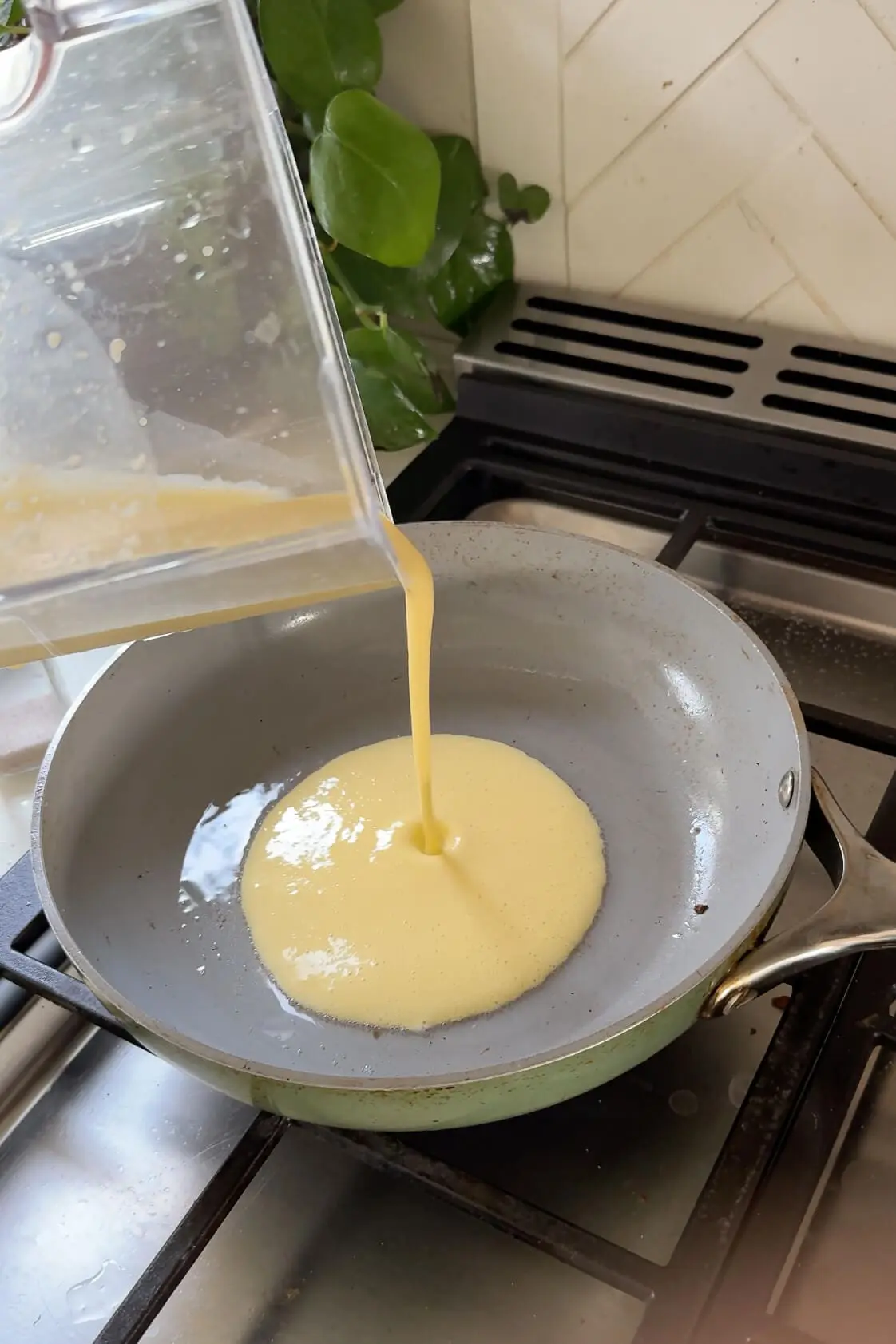 Blended cottage cheese and egg mixture being poured into a preheated skillet with a smooth gray surface, surrounded by a modern kitchen background.